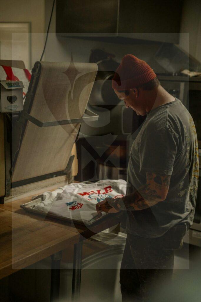 Photo by James Collington Adult man printing a custom t-shirt using a heat press in a dimly lit workshop.